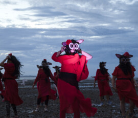 A dancer in a bush baby creature mask lit red, with her troupe behind her on the mudflats at dusk