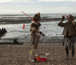 Two people dressed as cats, one dangling a toy fish, beside a red box on the shingle at low tide