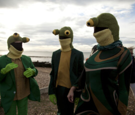 Three people in crocheted frog hoods talk on Whitstable beach
