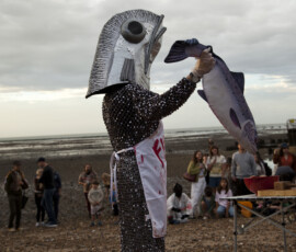 A performer in a giant fish head holds up a smaller fish, eye to eye