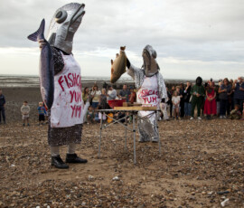 Two performers in giant fish heads and aprons hold up fish in front of a watching crowd