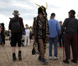 A musician in a goat mask carries a clarinet through the crowd