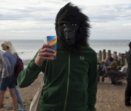 A person in a gorilla mask and green tracksuit raises a cup on the beach