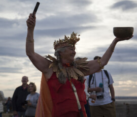 A woman in a gold crown and orange robe raises a singing bowl on Whitstable beach as people watch