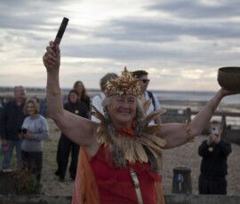 A smiling woman in a gold crown and feathers raises a singing bowl on Whitstable beach