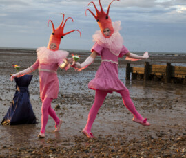 Two dancers in prawn headdresses leap across the wet mudflats at low tide