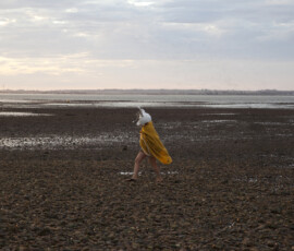 A figure in a white rabbit mask and yellow blanket walks across the mudflats at low tide