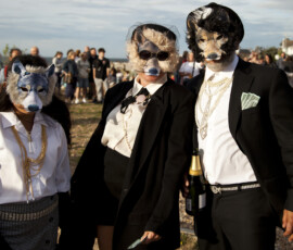 Three people in wolf masks, dark suits and pearls pose on Whitstable beach